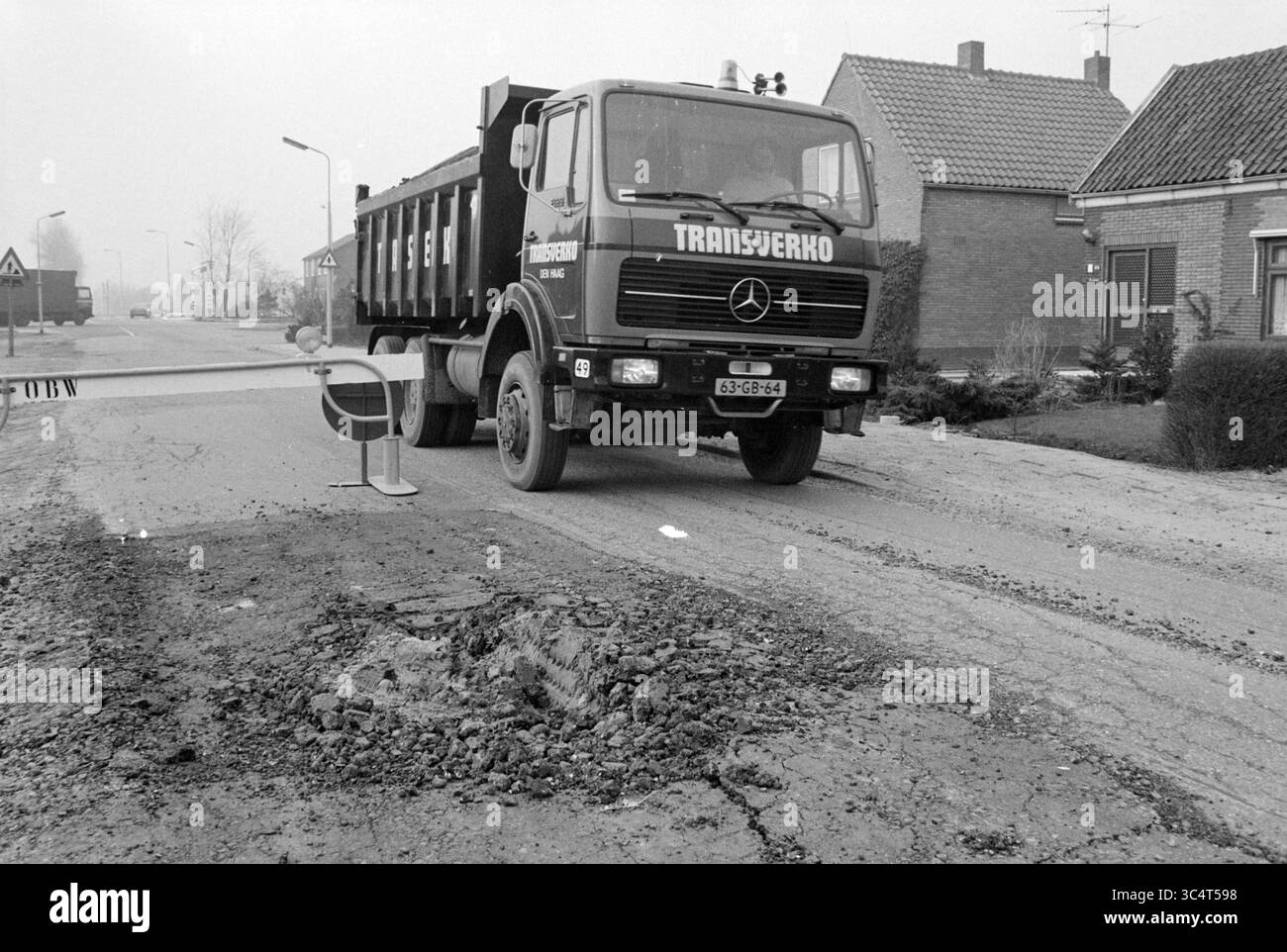 Buchi nel fondo stradale Rijnlanderweg Hoofddorp, strade, costruzione di strade, piantagione di strade, Hoofddorp, Rijnlanderweg, Paesi Bassi, 20-03-1979 Whizgle News, Dutch Desk, Paesi Bassi, 1950-2000 Un autocarro per impieghi gravosi con vano di carico verde e nero è parcheggiato su una strada segnata da un'importante poppa, mentre nelle vicinanze si trova una barriera stradale che segnala i lavori stradali. Edifici residenziali fiancheggiano lo sfondo, avvolti in un'atmosfera nebbiosa. Foto Stock