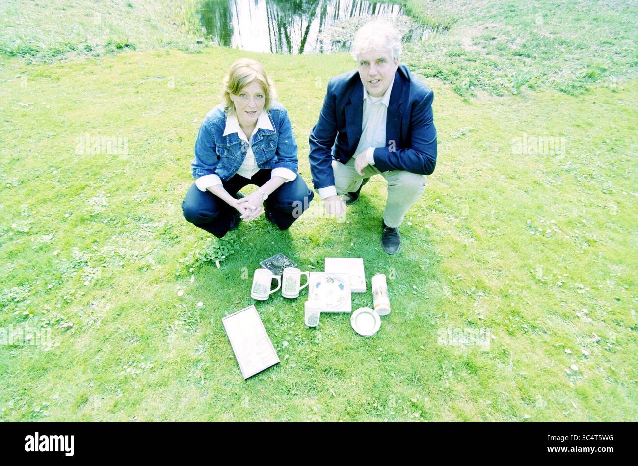 Coppia (?) Mostra materiale promozionale in vendita Whizgle News, Dutch Desk, Paesi Bassi, 1950-2000 un uomo e una donna si inginocchiano su un'area erbosa, circondata da vari oggetti disposti a terra, tra cui tazze e carte, con una tranquilla fontana visibile sullo sfondo. Foto Stock