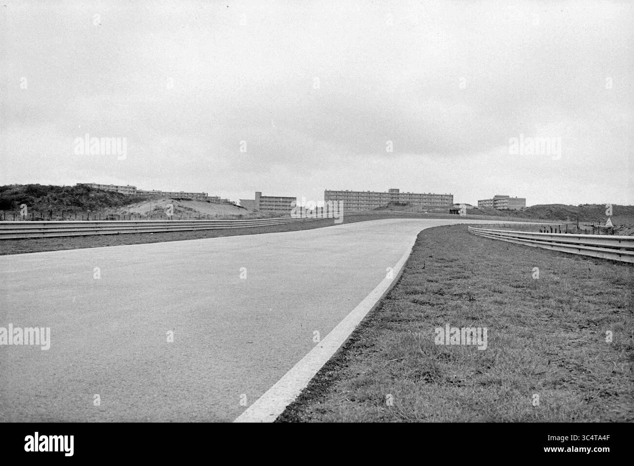 Appartamenti presso il circuito di Zandvoort, Circuit Zandvoort, esterno, 26-03-1975 Whizgle News, Dutch Desk, i Paesi Bassi, 1950-2000 Una strada tortuosa si estende in lontananza, fiancheggiata da banchi erbosi e bassi edifici che sorgono sullo sfondo sotto un cielo coperto. Foto Stock
