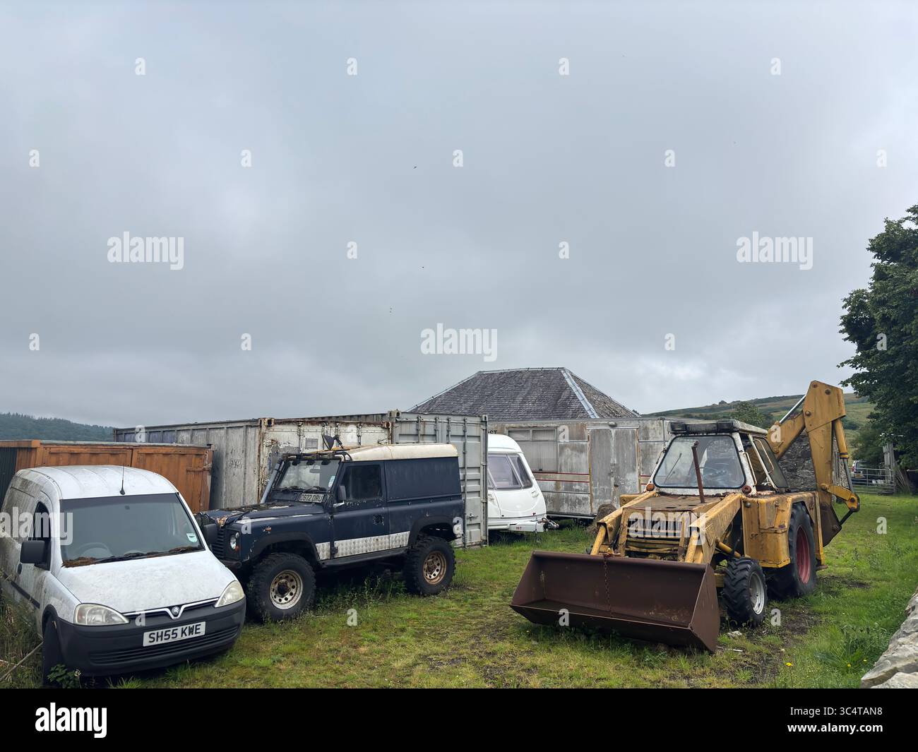 Auto abbandonate e un'arrugginente scavatrice sono rimaste in un campo nel Lothian occidentale, raffigurando il decadimento rurale, recuperando macchinari e veicoli sovradimensionati sotto lo sci estivo Foto Stock