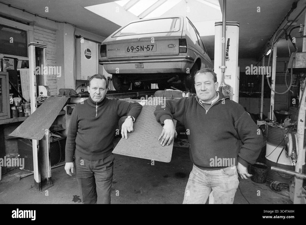 Azienda automobilistica Whizgle News, Dutch Desk, Paesi Bassi, 1950-2000 due uomini stanno in un garage, appoggiandosi casualmente contro un veicolo sollevato su un ascensore. Lo sfondo presenta strumenti e attrezzature tipici di un'officina di riparazione auto, mentre la luce solare filtra dall'alto, illuminando la scena. Foto Stock