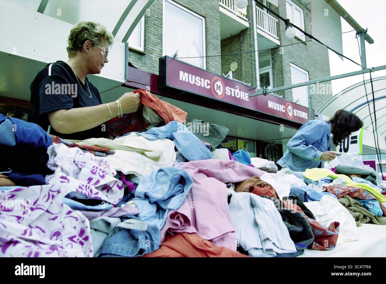 Midsummer, IJmuiden, IJmuiden, Paesi Bassi, 13-06-2002 Whizgle News, Dutch Desk, Paesi Bassi, 1950-2000 due persone curiosano tra un vibrante assortimento di vestiti impilati su un tavolo all'esterno di un negozio di musica, alla ricerca di gemme nascoste tra i vari tessuti e colori. Foto Stock