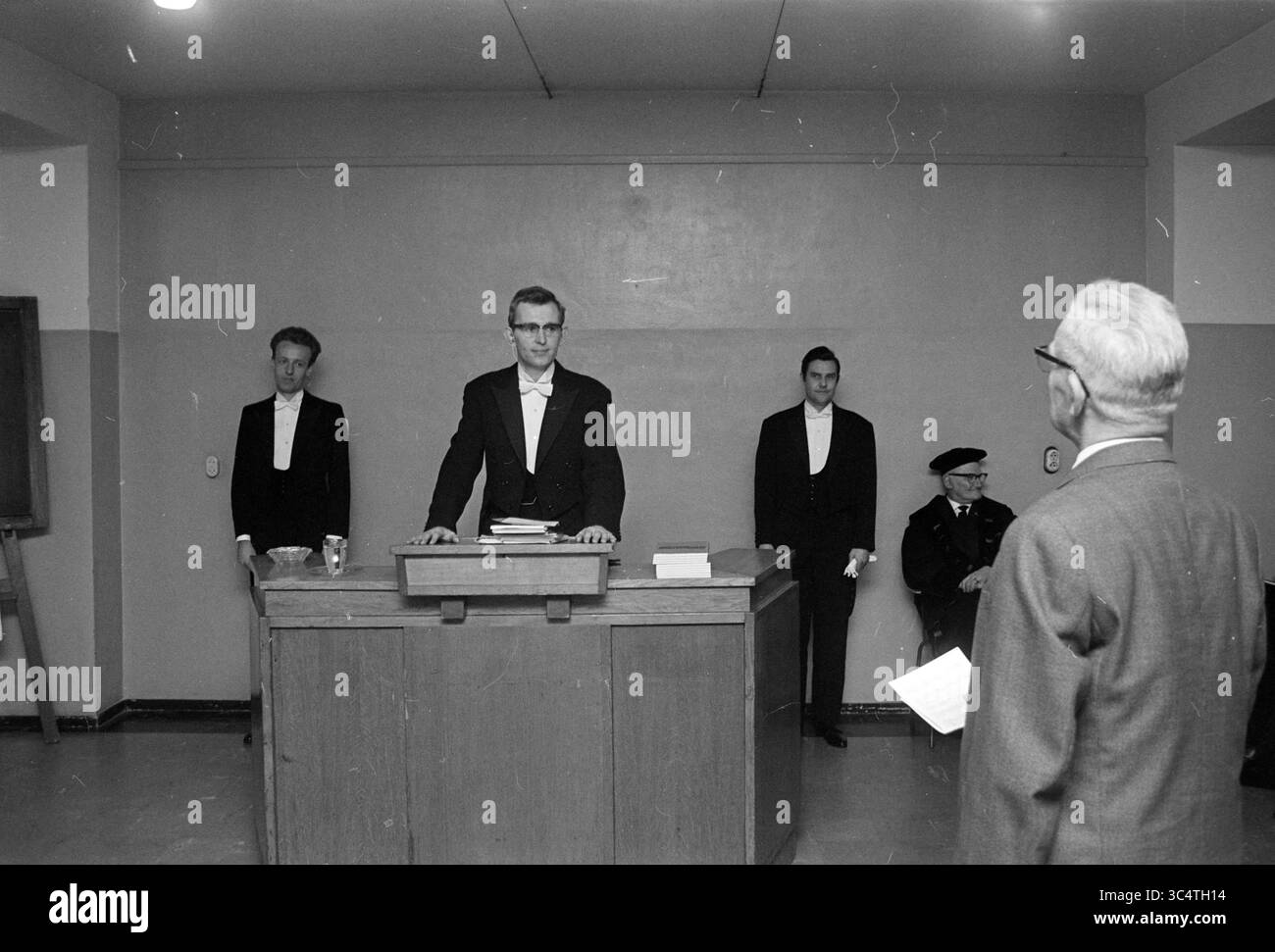 Presentazione della laurea a uno studente laureato in un Tuxedo Whizgle News, Dutch Desk, Paesi Bassi, 1950-2000 Un ambiente formale con una figura centrale su un podio, rivolgendosi a un pubblico. Due uomini stanno ai lati, mentre un signore più anziano ascolta attentamente dal fronte. L'atmosfera trasmette serietà e anticipazione, suggerendo una discussione o un evento significativo in corso. Foto Stock
