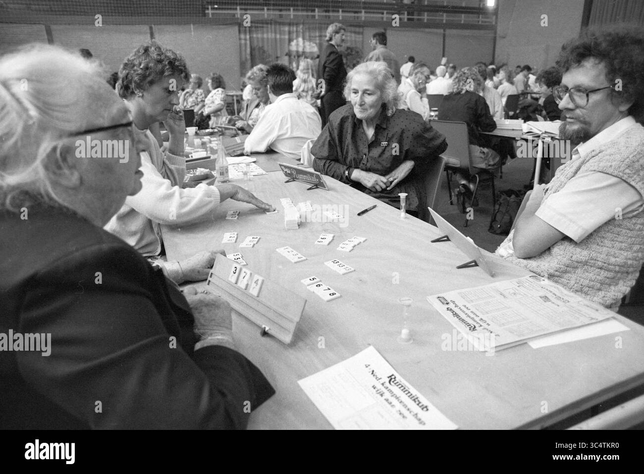 Dutch Rummikub Championships Wijk aan Zee, Giochi e giocattoli, competizioni, Wijk aan Zee, 26-08-1989 Whizgle News, Dutch Desk, Paesi Bassi, 1950-2000 Un gruppo di persone partecipa a un vivace gioco di carte in un tavolo affollato, con carte e punteggi distribuiti davanti a loro. I partecipanti sono concentrati sui loro turni, condividendo risate e conversazioni in un'atmosfera comune in un ambiente spazioso. Foto Stock