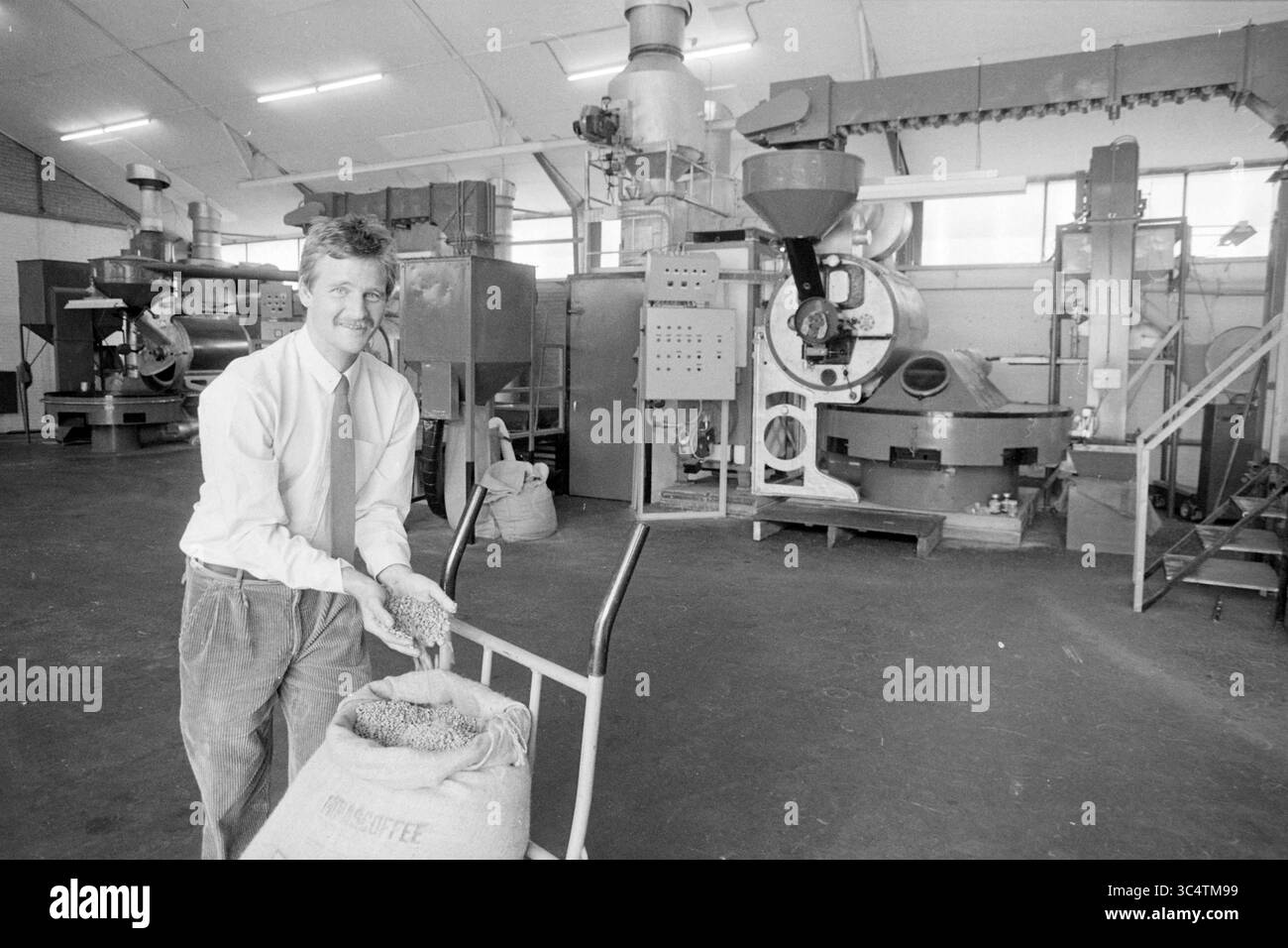 Tijs Sterrenberg della torrefazione di caffè de Amstel Boesingheliede., caffè, Boesingheliede, 01-08-1988 Whizgle News, Dutch Desk, i Paesi Bassi, 1950-2000 Un uomo si trova accanto a un carrello pieno di grani, sorridendo mentre espone il prodotto. In background, i macchinari industriali sono impegnati nella lavorazione dei cereali, evidenziando l'ambiente vivace di un impianto di lavorazione dei cereali. Foto Stock