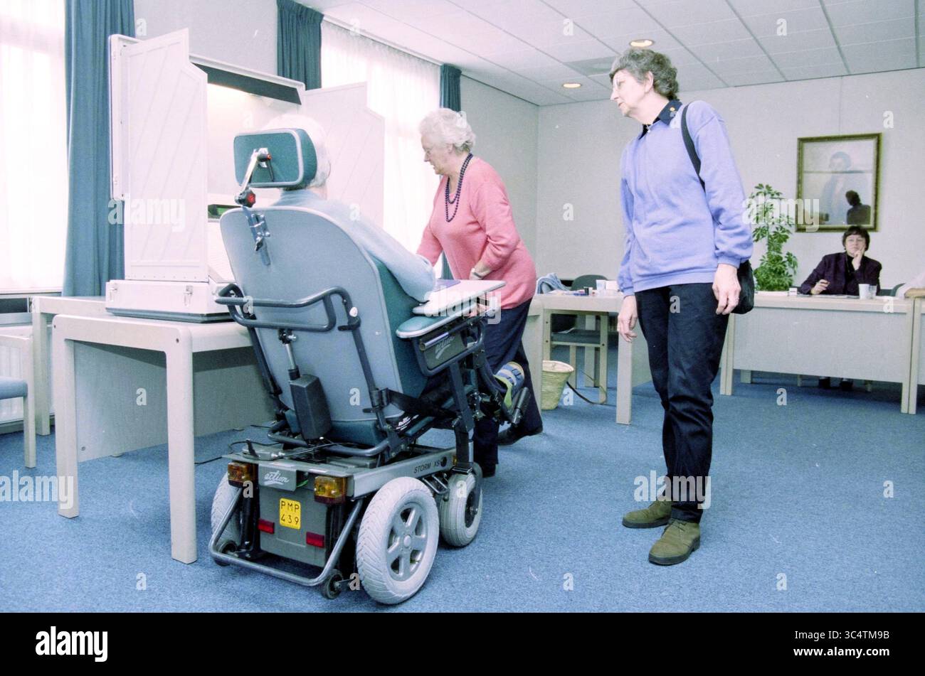 Vote Nieuw Unicum, Zandvoort, Zandvoort, 14-05-2002 Whizgle News, Dutch Desk, i Paesi Bassi, 1950-2000 una donna anziana in uno scooter mobile si avvicina a una scrivania con un ampio divisorio, mentre un'altra donna si trova nelle vicinanze, osservando l'interazione. L'ambiente sembra essere un'area d'attesa o un ufficio, caratterizzato da arredi semplici e da pochi individui impegnati in varie attività sullo sfondo. Foto Stock