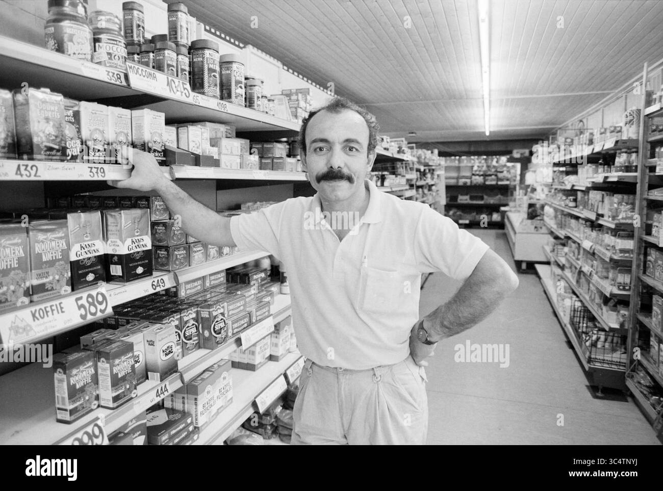 Moestafa Hansen supermarket Abbenes, supermercato, Abbenes, 21-08-1989 Whizgle News, Dutch Desk, i Paesi Bassi, 1950-2000 Un uomo si trova con fiducia nel corridoio di un negozio di alimentari, appoggiato su uno scaffale pieno di conserve e altri prodotti, mostrando un'espressione amichevole e una camicia ben stampata. Foto Stock