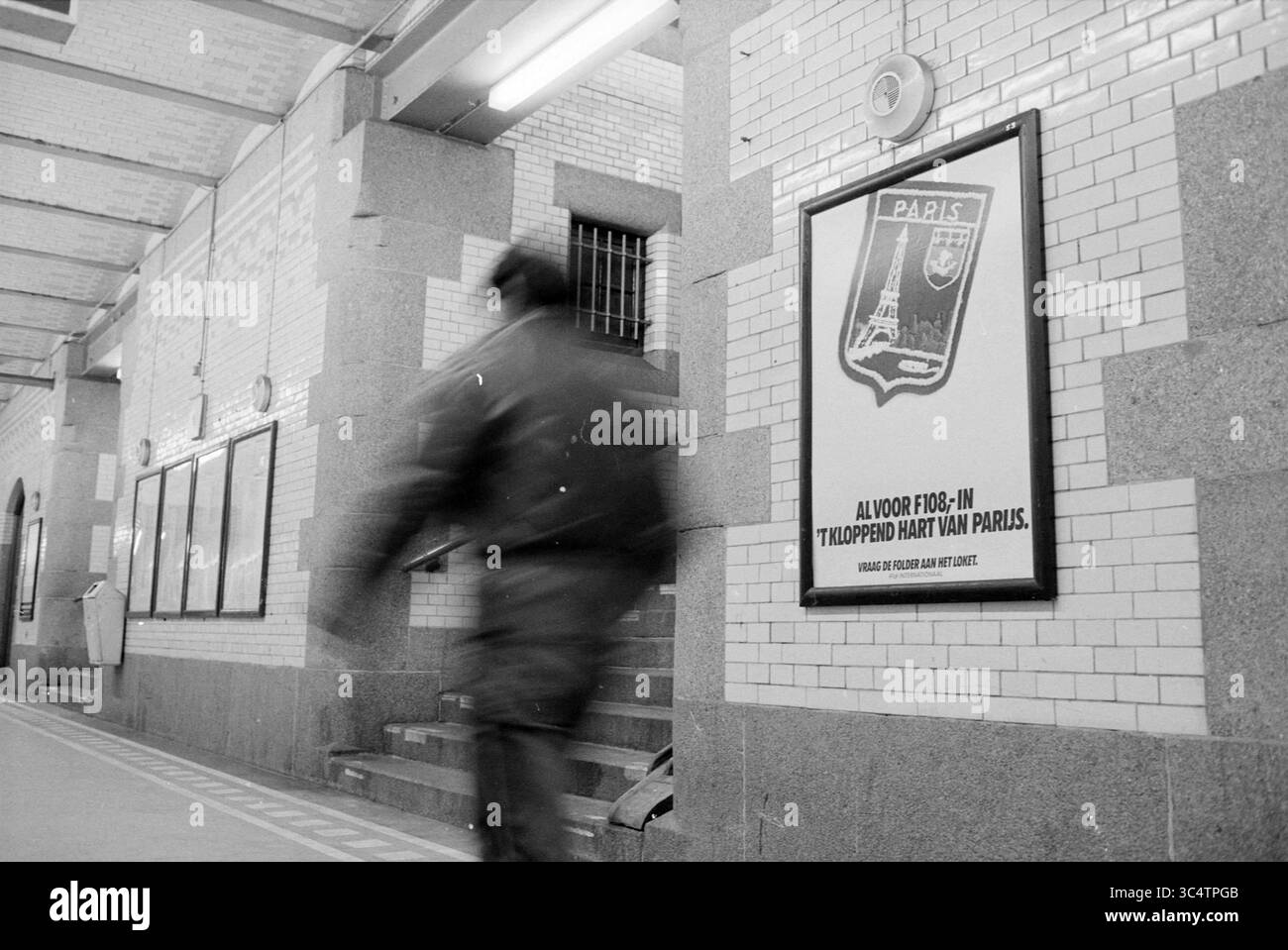 Dettaglio della stazione ferroviaria di Haarlem, Haarlem, The Netherlands Whizgle News, Dutch Desk, The Netherlands, 1950-2000 Una figura sfocata si precipita davanti a un poster di viaggio vintage per Parigi, con la Torre Eiffel, mentre si trova in una vivace stazione della metropolitana adornata da pareti piastrellate e una scala che conduce verso l'alto. Foto Stock