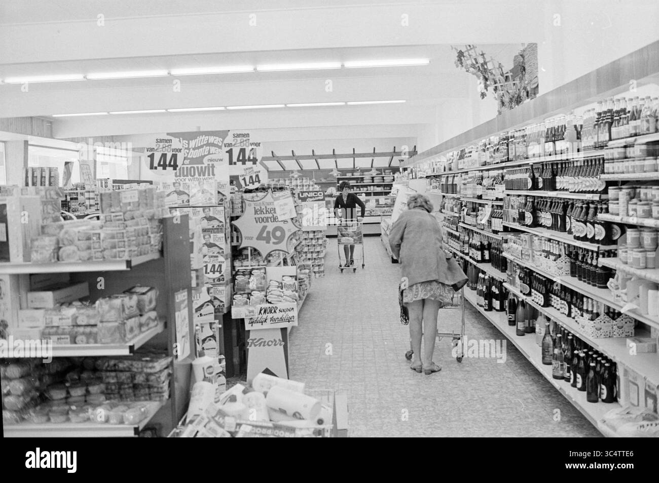 Int Supermercato, interni, 30-08-1971 Whizgle News, Dutch Desk, Paesi Bassi, 1950-2000 un corridoio in un negozio di alimentari pieno di scaffali pieni di vari prodotti, tra cui alimenti e bevande. Un acquirente spinge un carrello mentre un'altra persona naviga in lontananza, circondato da una serie di confezioni colorate e cartelli promozionali. Foto Stock