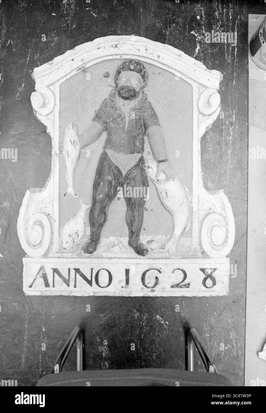 Facciata in pietra uomo con pesce, 1628, Beverwijk, facciata, pietre per facciate, Beverwijk, Paesi Bassi, 01-02-1984 Whizgle News, Dutch Desk, Paesi Bassi, 1950-2000 Una scultura storica raffigurante una figura con due pesci, con l'anno '1628' iscritto sotto. La figura è ricoperta di abiti che suggeriscono un tema marittimo, sottolineando l'importanza della pesca in quell'epoca. L'opera d'arte è incorniciata da elementi decorativi che aggiungono fascino vintage. Foto Stock
