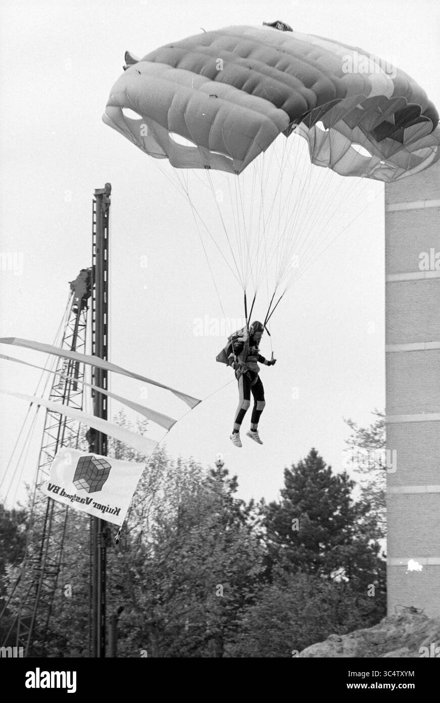 Primo polo Heemhaven, Heemstede (paracadutista), Heemstede, Paesi Bassi, 18-05-1994 Whizgle News, Dutch Desk, Paesi Bassi, 1950-2000 Un paracadutista scende con grazia, imbracato ed equipaggiato per un atterraggio, con una grande bandiera che sventola sotto e attrezzature da costruzione sullo sfondo. Gli alberi e una struttura urbana aggiungono contesto alla scena. Foto Stock