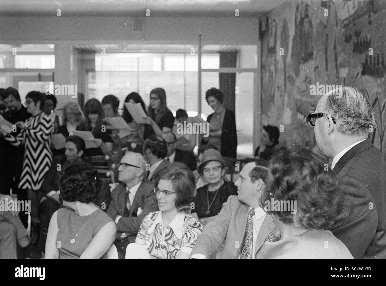 Anniversario signor la Blance, gemma. Library IJmuiden, Jubilee, anniversari, 21-04-1971 Whizgle News, Dutch Desk, i Paesi Bassi, 1950-2000 Un raduno di individui in un ambiente vivace, alcuni prendono appunti o leggono da fogli, mentre altri si dedicano alla conversazione. L'atmosfera è piena di attesa e di senso di comunità, dato che persone di varie età sono sedute a stretto contatto, con vivaci murales che adornano le pareti dietro di loro. Foto Stock