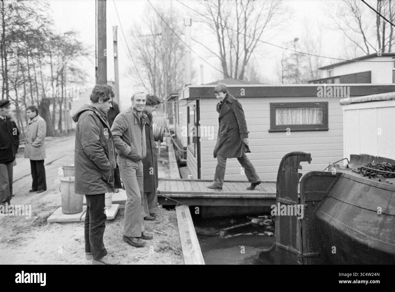 Alderman Kaptein Visit Waarderhaven, Aldermen, 20-12-1978 Whizgle News, Dutch Desk, Paesi Bassi, 1950-2000 Un gruppo di individui si trova vicino a una barca ormeggiata, interagendo casualmente mentre una persona cammina attraverso uno stretto ponte verso una casa galleggiante. Gli alberi allineano lo sfondo, a indicare un'impostazione tranquilla. Foto Stock
