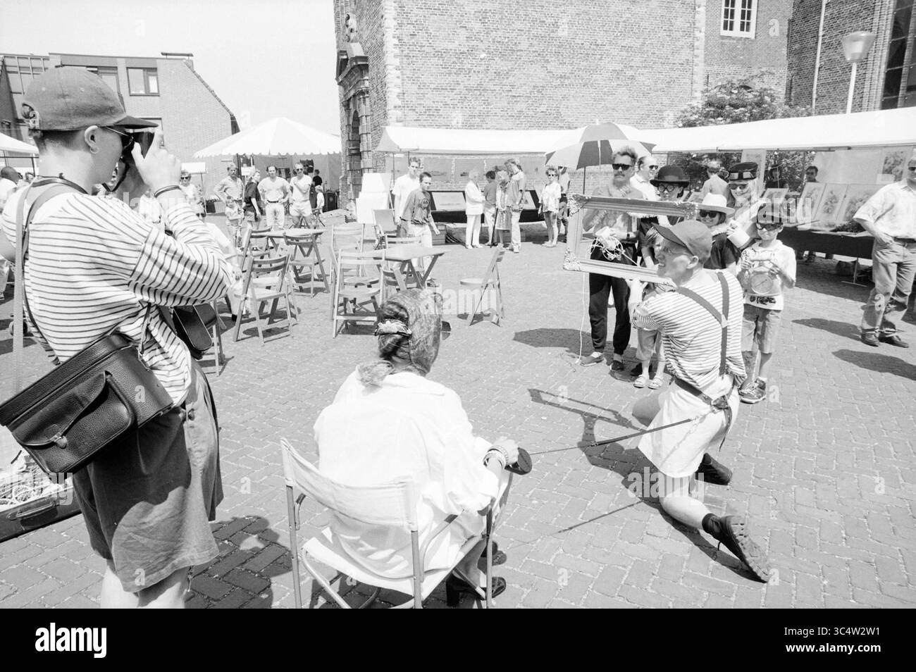 Art Market at Wijkertoren, B'wijk, Beverwijk, Paesi Bassi, 27-05-1992 Whizgle News, Dutch Desk, Paesi Bassi, 1950-2000 Una vivace scena di mercato all'aperto presenta un gruppo di persone impegnate in varie attività. Un bambino sta giocando a posare con un fiocco e una freccia mentre indossa abiti leggeri, catturando l'attenzione degli spettatori. Nelle vicinanze, una serie di bancarelle espone opere d'arte, e gli individui sono visti navigare e interagire in un'atmosfera allegra. Foto Stock