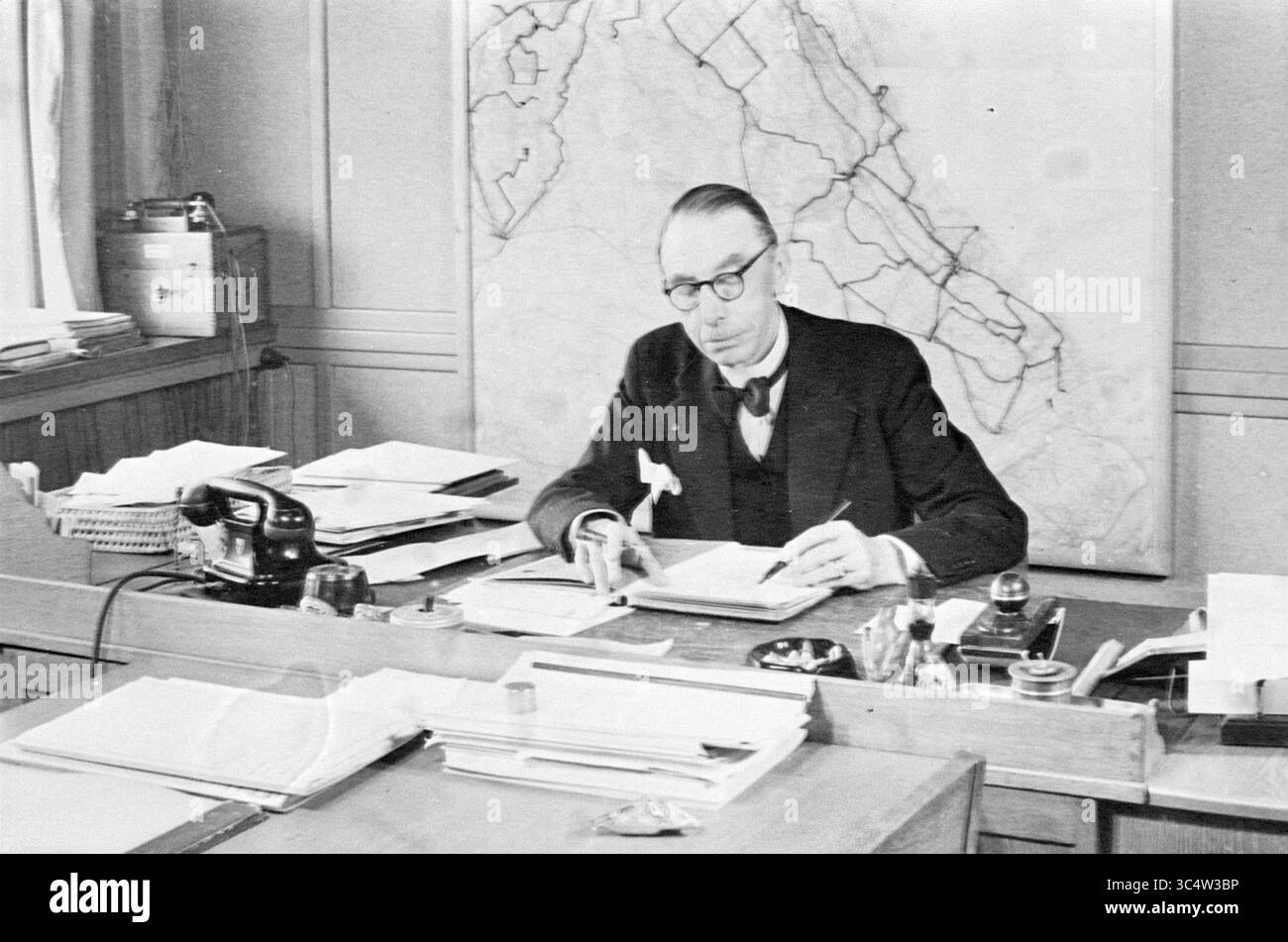 Anniversario HR.Hetem, Noord Zuid Hollandse Vervoersmaatschappij N.V., NZ, 23-07-1963','24-07-1963 Whizgle News, Dutch Desk, Paesi Bassi, 1950-2000 Un uomo siede su una scrivania disordinata, circondato da pile di documenti e documenti, mentre si concentra sulla scrittura di appunti. Sullo sfondo, una mappa di grandi dimensioni è fissata al muro, indicando una connessione con la pianificazione o l'organizzazione. Indossa una tuta formale e occhiali, proiettando un'aria di professionalità e concentrazione. Foto Stock