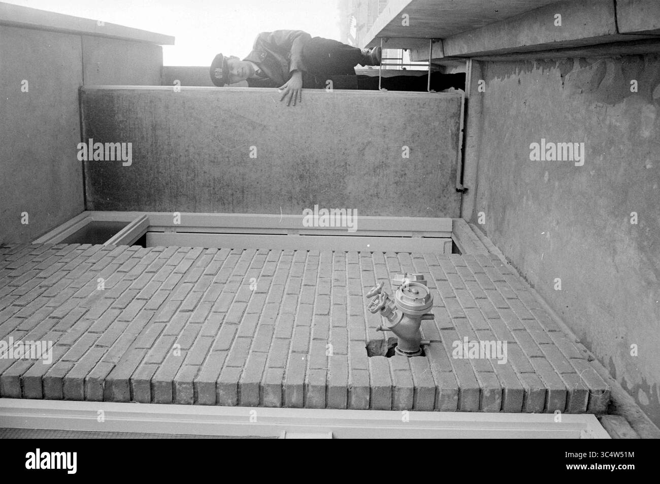 Strutture dei vigili del fuoco in appartamento a Schalkwijk, incendi, vigili del fuoco, costruzione di case e case, 20-04-1964 Whizgle News, Dutch Desk, Paesi Bassi, 1950-2000 Una figura si appoggia su una sporgenza, scorrendo verso il basso su una superficie di mattoni sottostante, dove un idrante d'acqua è parzialmente esposto attraverso un'apertura. Le pareti circostanti mostrano un aspetto testurizzato e non finito, che trasmette un ambiente industriale o urbano. Foto Stock