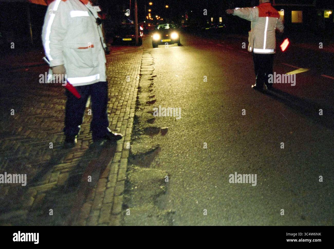 Controllo dell'alcool IJmuiden, IJmuiden, Paesi Bassi, 14-02-2002 Whizgle News, Dutch Desk, i Paesi Bassi, 1950-2000 due persone in giacche riflettenti dirigono il traffico di notte, una segnalando con una bacchetta di segnalazione mentre si trovano vicino a marciapiedi irregolari. Un veicolo si avvicina sullo sfondo, illuminato da lampioni. Foto Stock