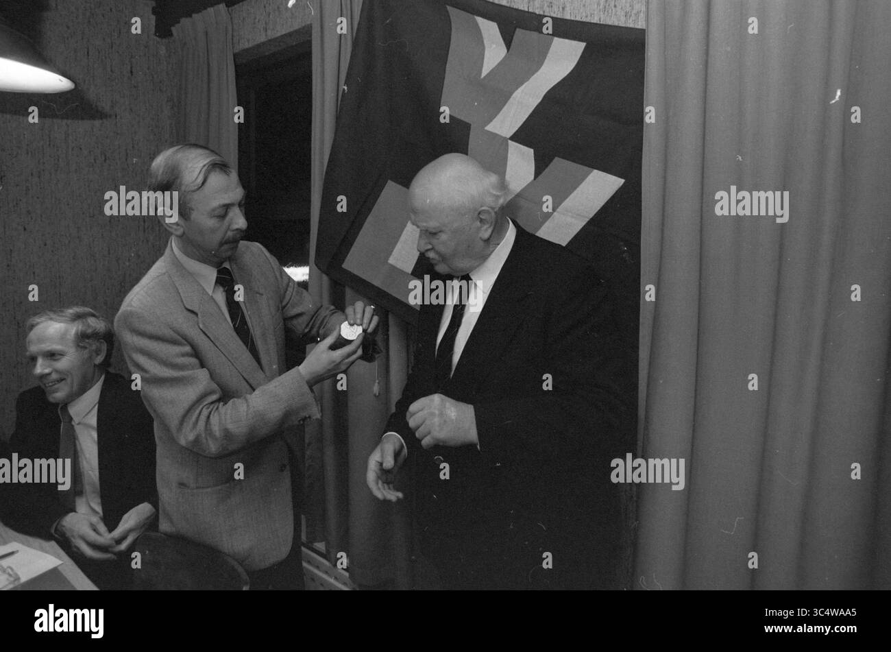 Lode Arie Klomp VVD Beverwijk, lode, Beverwijk, Paesi Bassi, 11-11-1987 Whizgle News, Dutch Desk, Paesi Bassi, 1950-2000 Un gruppo di uomini è impegnato in un momento formale, con uno che presenta un piccolo oggetto a un altro, che sembra riceverlo. Una bandiera con un distintivo emblema è appesa sullo sfondo, aggiungendo un contesto storico alla scena. Foto Stock