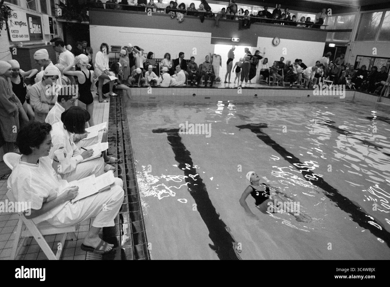 Competizione di nuoto Whizgle News, Dutch Desk, Paesi Bassi, 1950-2000 Una competizione di nuoto sincronizzata si svolge in un vivace centro acquatico, con i giudici che guardano con attenzione dal bordo. I concorrenti, tra cui un nuotatore che scivola attraverso l'acqua con un costume da bagno impressionante, mostrano le loro abilità in mezzo a un pubblico pieno di attesa ed eccitazione. Foto Stock
