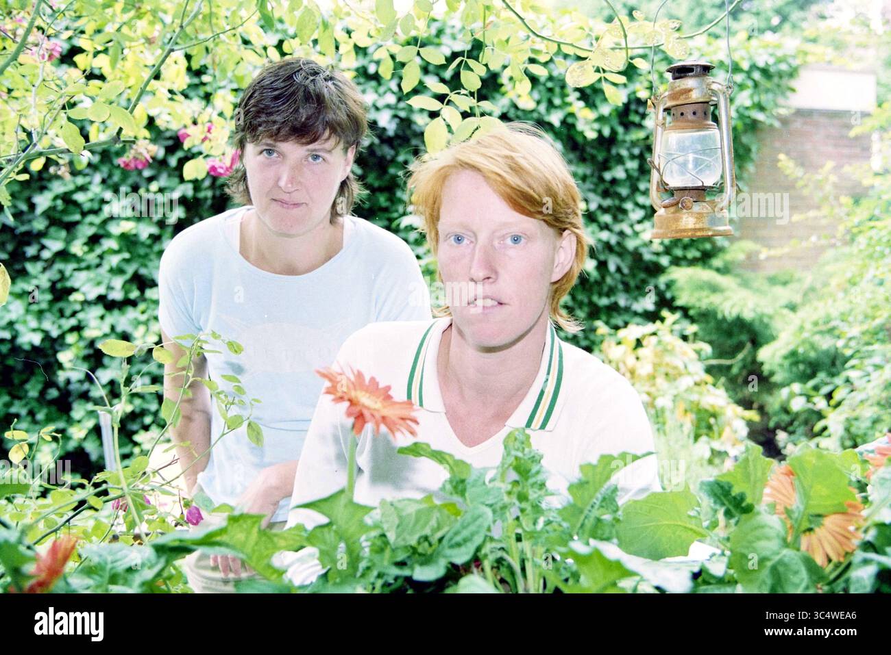 Gruppo teatrale de Polder, 05-07-1999 Whizgle News, Dutch Desk, Paesi Bassi, 1950-2000 due persone si stagliano tra vegetazione lussureggiante e fiori vivaci, con una lanterna d'epoca sospesa nelle vicinanze. Una persona guarda attentamente verso lo spettatore, mentre l'altra sembra impegnata nella bellezza circostante del giardino. Foto Stock