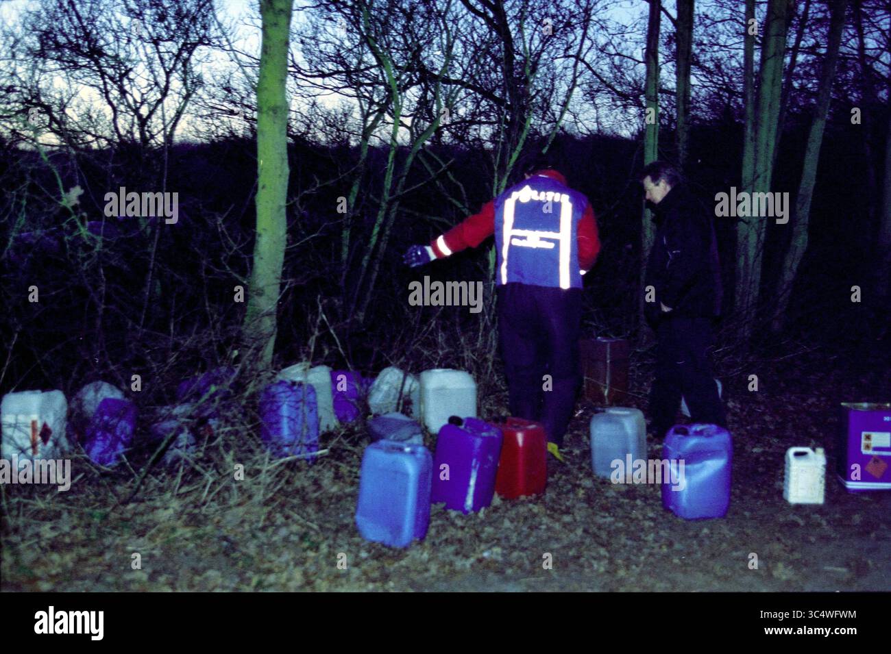 Ecstasy waste, Beeckestijn, 09-12-2002 Whizgle News, Dutch Desk, Paesi Bassi, 1950-2000 due persone si trovano vicino a una collezione di taniche allineate lungo un'area boscosa, con una persona che punta verso le lattine in mezzo alla luce che svanisce. La scena suggerisce un contesto serio o investigativo. Foto Stock