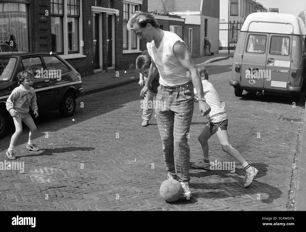 Hans Werdekker Street football da Costastraat, calciatori e allenatori, 06-05-1988 Whizgle News, Dutch Desk, Paesi Bassi, 1950-2000 bambini giocano in strada mentre un uomo dribbla abilmente un pallone da calcio, creando una scena vivace piena di gioia ed energia in una giornata di sole. Le auto d'epoca fiancheggiano la strada acciottolata, aggiungendo un tocco di nostalgia al momento giocoso. Foto Stock