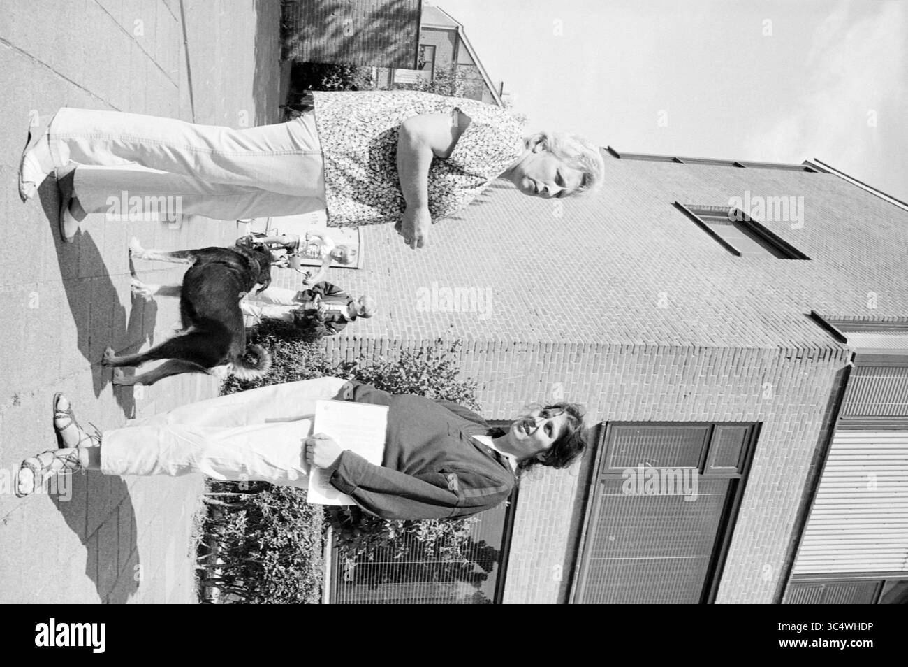 Cattive case Schalkwijk fam. Wandelee, costruzione di case e case, 27-06-1984 Whizgle News, Dutch Desk, Paesi Bassi, 1950-2000 due donne si trovano all'aperto, impegnate in conversazione, mentre un cane è nelle vicinanze. Sullo sfondo, una persona sta andando in bicicletta, e un edificio con grandi finestre si infila lateralmente. L'ambiente sembra informale e orientato alla comunità. Foto Stock