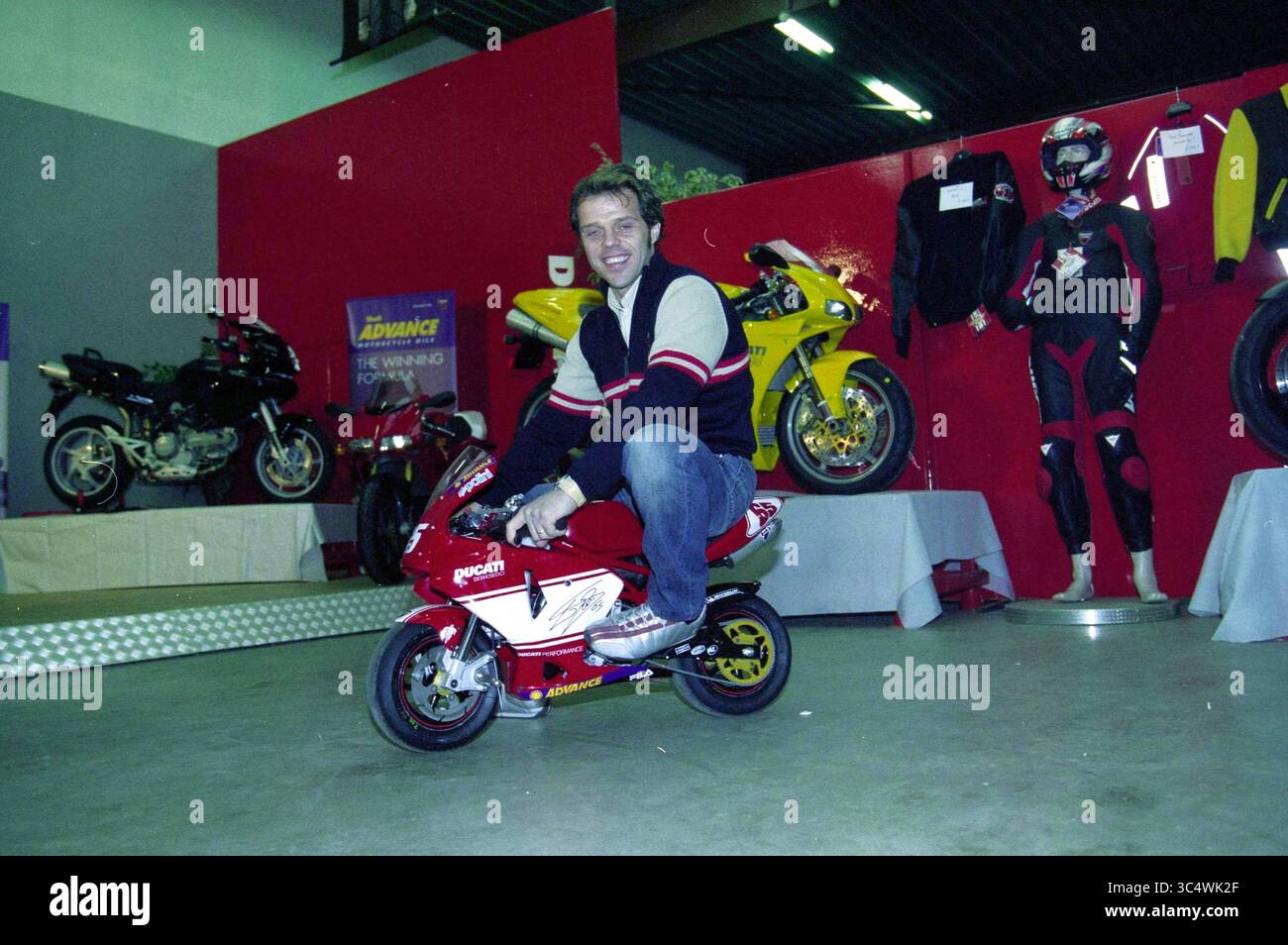 Apertura del negozio di motociclette Nieuw-Vennep, Loris Capirossi, Nieuw-Vennep, 12-12-2003 Whizgle News, Dutch Desk, i Paesi Bassi, 1950-2000 Un uomo posa giocosamente su una moto Ducati in miniatura, circondato da una mostra di moto più grandi e di equipaggiamento da corsa. L'ambiente vivace mette in risalto l'emozione della cultura motociclistica. Foto Stock
