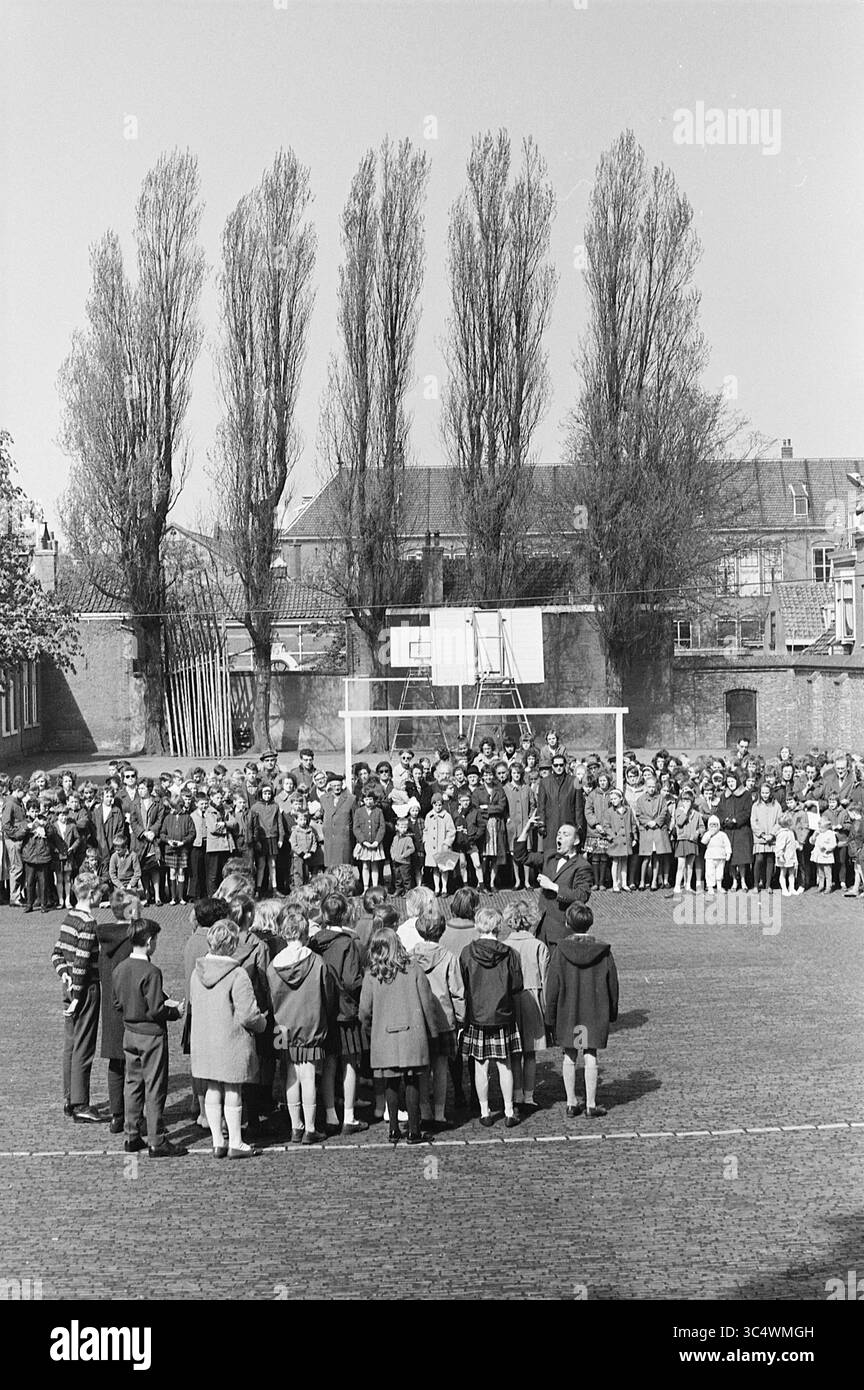 Coro per bambini con pubblico all'esterno, 05-05-1964 Whizgle News, Dutch Desk, Paesi Bassi, 1950-2000 Un grande gruppo di scolari si riunisce in un parco giochi, con alcuni in piedi in cerchio mentre altri guardano dai lati. Alberi alti fiancheggiano lo sfondo e un canestro da basket si erge in modo prominente. La scena cattura un vivace evento scolastico all'aperto, pieno di energia giovanile e cameratismo. Foto Stock