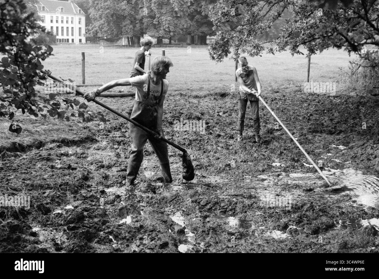 Dredge Pond estate Waterland, pulizia, stagni, Velsen, 04-09-1987 Whizgle News, Dutch Desk, Paesi Bassi, 1950-2000 tre persone sono impegnate nel lavoro manuale, utilizzando strumenti a lunga movimentazione per lavorare in un'area fangosa. L'impostazione presenta un campo erboso e alberi sullo sfondo, con un edificio visibile in lontananza. L'attenzione è concentrata sui loro sforzi per gestire il terreno impegnativo. Foto Stock