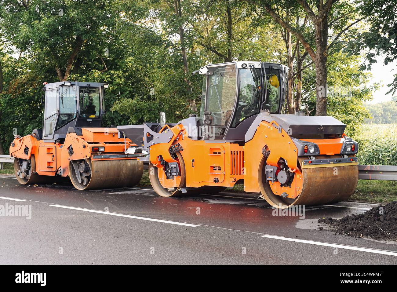 Rulli su asfalto. Macchine per l'edilizia in loco. Attrezzature pesanti vicino alla foresta. Costruzione di strade industriali. Due compattatori arancioni. Foto Stock