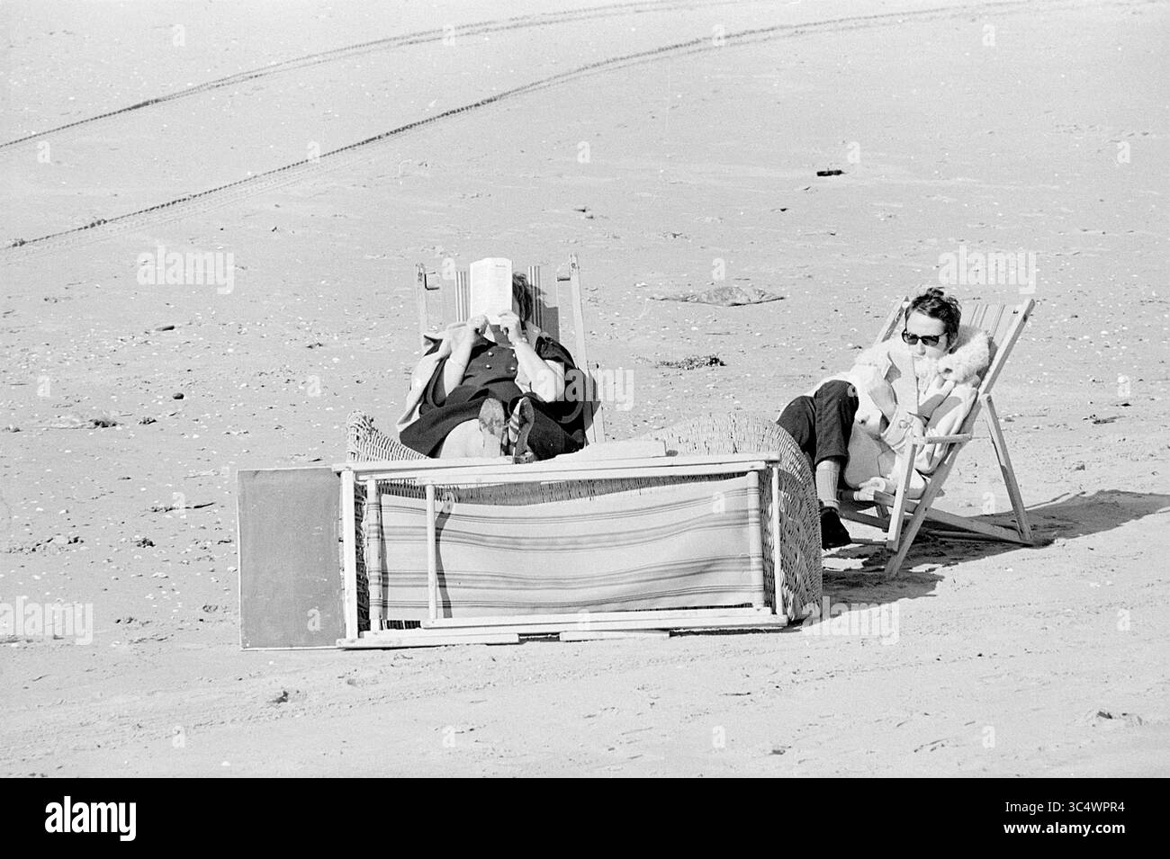 Persone che prendono il sole sulla spiaggia, sulla spiaggia e sulle spiagge, 30-09-1969 Whizgle News, Dutch Desk, Paesi Bassi, 1950-2000 due persone si rilassano sulla spiaggia, una immersa in un libro, mentre l'altra si siede tranquillamente al loro fianco. Una coperta intrecciata avvolge le sedie e la tranquilla costa si estende dietro di esse. Foto Stock