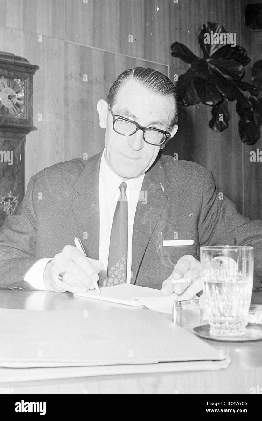 Consiglieri municipali di Beverwijk, Consiglio municipale di Beverwijk, 21-12-1970 Whizgle News, Dutch Desk, Paesi Bassi, 1950-2000 Un uomo ben vestito siede ad una scrivania, concentrandosi sulla scrittura su un foglio di carta. Indossa occhiali e una cravatta, e un bicchiere d'acqua riposa nelle vicinanze, accanto a un posacenere. Una grande pianta è visibile sullo sfondo, aggiungendo un tocco di vita all'ambiente dell'ufficio. Foto Stock