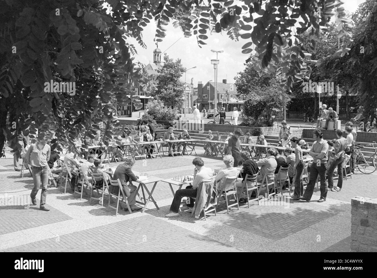 Scacchi simultanei all'aperto Nielenplein Heemskerk, scacchi, Heemskerk, Burgemeester Nielenplein, 18-07-1981 Whizgle News, Dutch Desk, Paesi Bassi, 1950-2000 Un vivace raduno all'aperto si svolge in un parco, dove le persone sono impegnate in varie attività presso una serie di tavoli. Alcuni sono concentrati sui giochi, mentre altri chiacchierano o si rilassano. Gli alberi lussureggianti forniscono ombra e uno sfondo di edifici accenna ad un invitante ambiente urbano. Le biciclette sono parcheggiate nelle vicinanze, aggiungendo un'atmosfera informale. Foto Stock