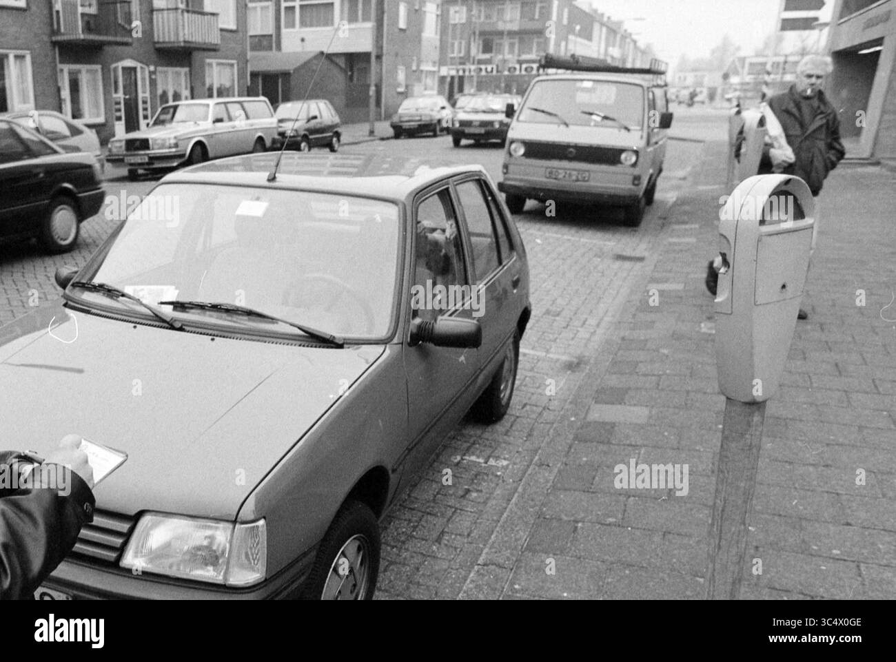 Addetto al parcheggio in azione, IJmuiden, Paesi Bassi, 10-02-1993 Whizgle News, Dutch Desk, i Paesi Bassi, 1950-2000 Una piccola auto parcheggiata lungo una strada costeggiata da vari veicoli, mentre una persona sembra interagire con essa. Nelle vicinanze, un parcheggio si trova accanto al marciapiede e un pedone osserva da lontano. La scena cattura un momento della vita quotidiana in città. Foto Stock