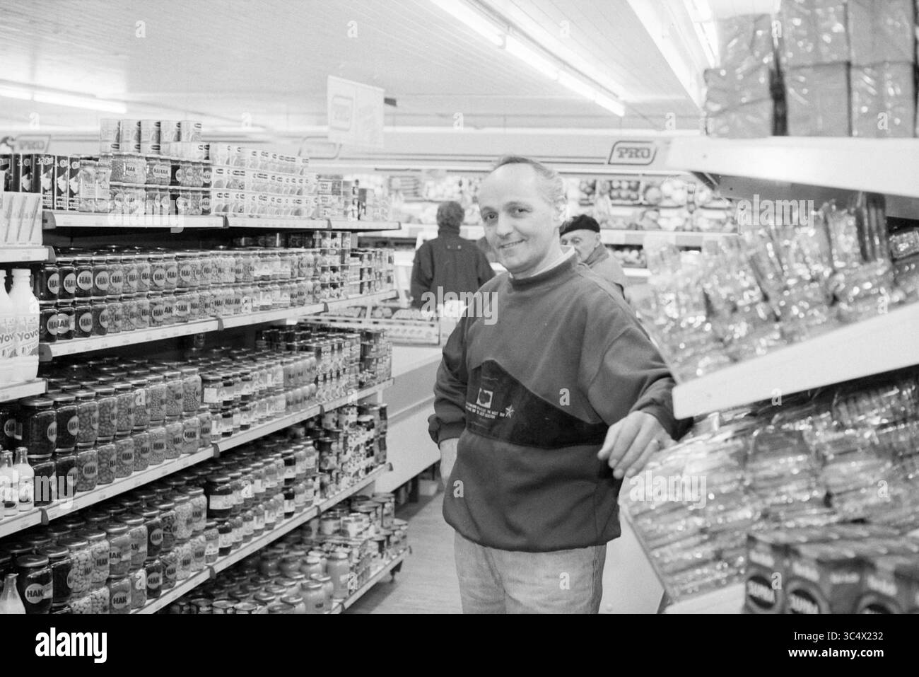 Chris van Haastert, Abbenes, Abbenes, 15-01-1992 Whizgle News, Dutch Desk, i Paesi Bassi, 1950-2000 Un uomo si trova in una corsia di un negozio di alimentari, circondato da scaffali pieni di conserve e snack, mentre un'altra persona si vede sullo sfondo mentre sfoglia i prodotti. La scena cattura un momento della vita quotidiana dello shopping. Foto Stock