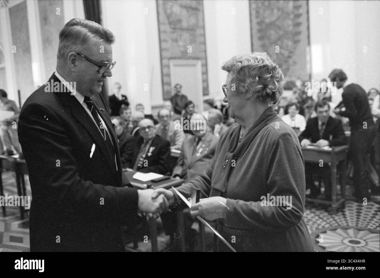 Presentazione delle croci di resistenza da parte del Commissario della Regina Roel de WIT, nella Casa Provinciale, Prov. Stati, Prov. Gestione dell'acqua, Provincial House, Haarlem, Paesi Bassi, 21-01-1982 Whizgle News, Dutch Desk, Paesi Bassi, 1950-2000 Un evento formale in cui un uomo e una donna sono impegnati in una stretta di mano, simboleggiando uno scambio o un riconoscimento. Sullo sfondo, un gruppo di persone è riunito, osservando l'interazione, creando un senso di comunità e impegno in un ambiente cerimoniale. Foto Stock