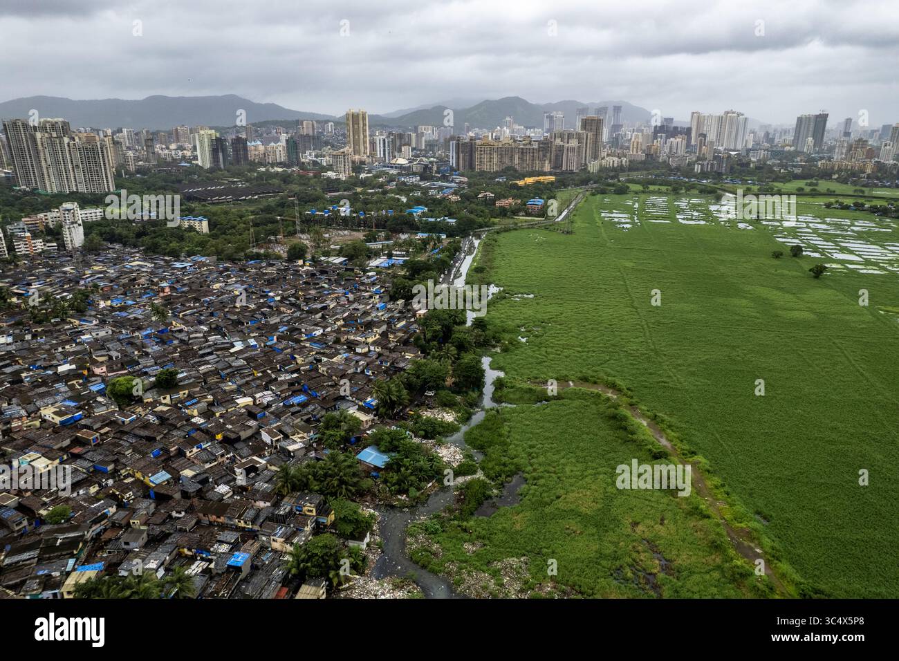 Vista aerea di forti contrasti tra fitte baraccopoli urbane e un vivace campo verde, diviso da un tortuoso fiume, Mumbai, Maharashtra, India. Foto Stock