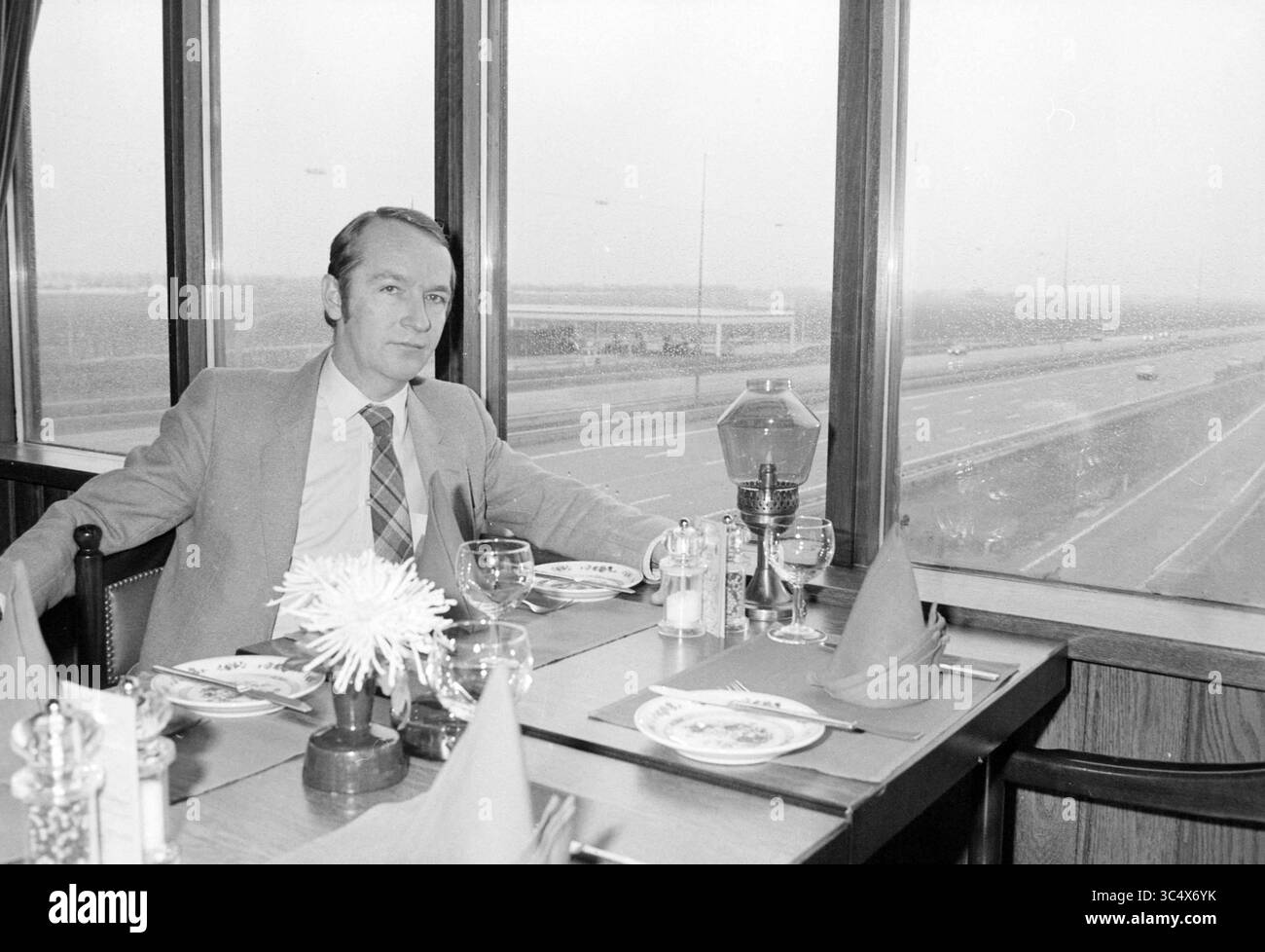 Direttore De Leeuw Ricks, ristorante lungo la strada Hoofddorp, Persons catering, Hoofddorp, Paesi Bassi, 31-01-1983 Whizgle News, Dutch Desk, Paesi Bassi, 1950-2000 Un gentiluomo siede ad un tavolo da pranzo per un pasto, con piatti e utensili ben disposti. Guarda attentamente fuori da una grande finestra che offre una vista su un'autostrada trafficata, mentre un pezzo centrale decorativo di fiori e condimenti adornano il tavolo intorno a lui. Foto Stock