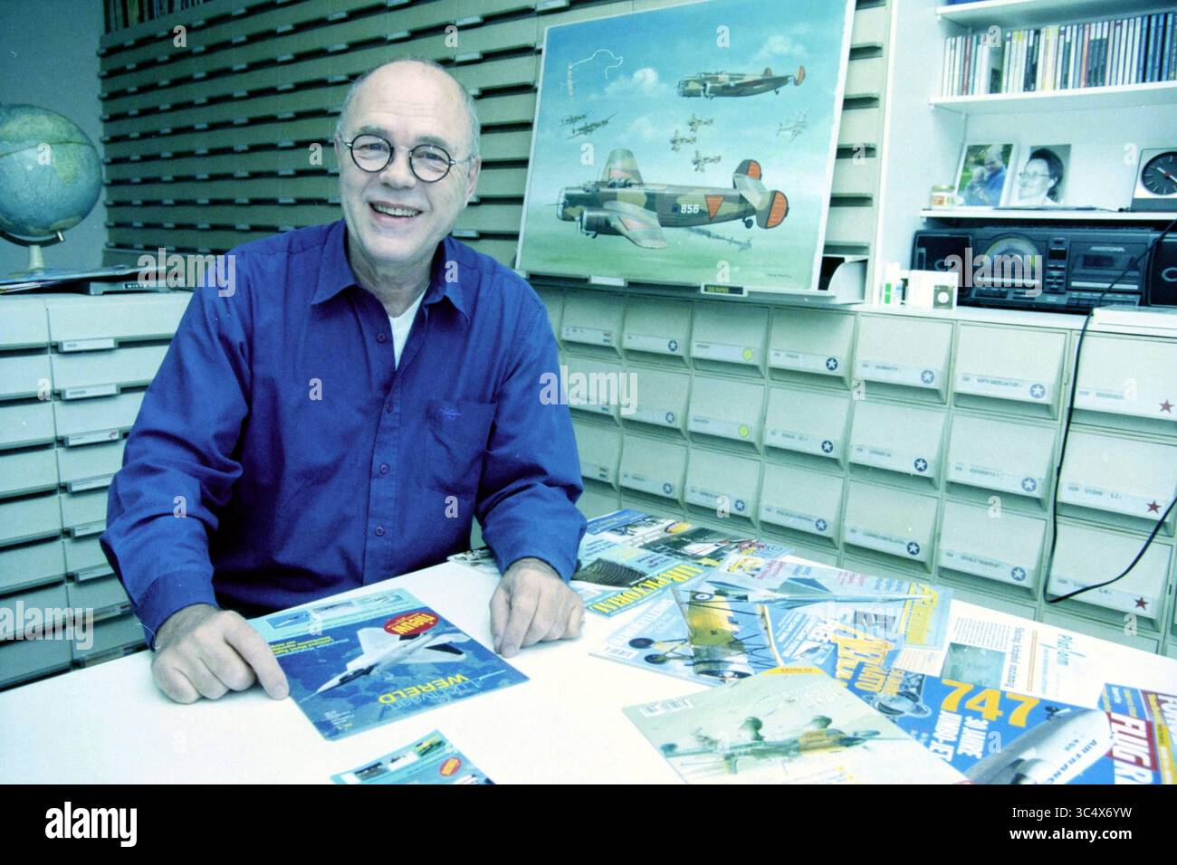 Thijs Postma, Hoofddorp, Paesi Bassi, 11-04-1999 Whizgle News, Dutch Desk, i Paesi Bassi, 1950-2000 Un individuo allegro con gli occhiali siede su un tavolo coperto da varie riviste di aviazione e opere d'arte di aerei, circondato da un contesto ben organizzato. L'atmosfera trasmette la passione per l'aviazione e la storia. Foto Stock