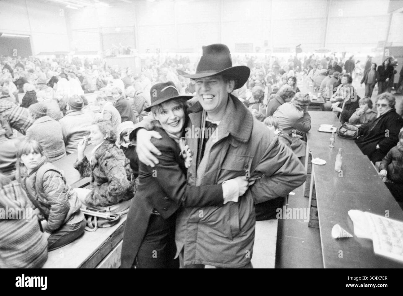 Report Black Market Beverwijk, Market, 00-00-1981 Whizgle News, Dutch Desk, Paesi Bassi, 1950-2000 Una coppia sorridente si abbraccia in una folla vivace, entrambi vestiti con eleganti capospalla e cappelli, trasudando un senso di gioia e connessione in un evento movimentato. Lo sfondo è pieno di partecipanti, tutti impegnati in vivaci conversazioni e attività. Foto Stock