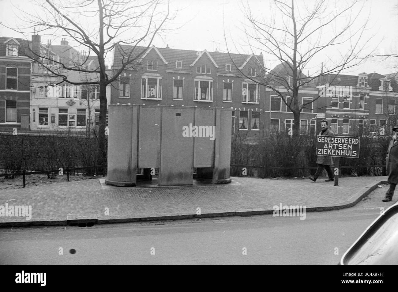 Cartello a Urinal, cartelli, 20-02-1964 Whizgle News, Dutch Desk, Paesi Bassi, 1950-2000 Una scena di strada presenta una struttura simile a un bagno pubblico, fiancheggiato da alberi, con una persona con un cartello con la scritta "Gereserveerd Artsen Ziekenhuis", che indica lo spazio riservato ai medici in un ospedale. Dietro, edifici tradizionali fiancheggiano la strada, contribuendo all'atmosfera urbana. Foto Stock