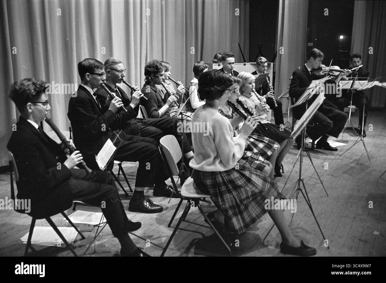 Scuola orchestra Pr.Bernhard H.B.S. IJmuiden, musica, 22-02-1963 Whizgle News, Dutch Desk, Paesi Bassi, 1950-2000 Un gruppo di giovani musicisti siede in formazione, suonando vari strumenti a fiato. Sono concentrati e impegnati, vestiti con abbigliamento formale, mentre si esibiscono su un palcoscenico illuminato da luci soffuse. Gli stand musicali tengono i loro spartiti, contribuendo all'atmosfera di un concerto o di un recital. Foto Stock