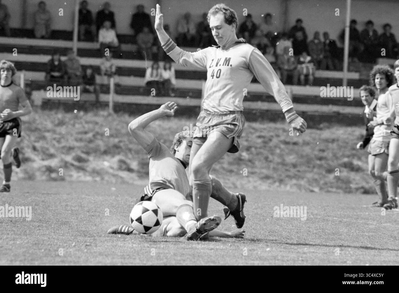 Zandvoort Meeuwen - Volendam, Football Zandvoortse Meeuwen Halfway, 25-04-1982 Whizgle News, Dutch Desk, Paesi Bassi, 1950-2000 due giocatori di calcio sono impegnati in una partita riscaldata sul campo. Un giocatore, vestito con un'uniforme di colore chiaro, sta scivolando nel tentativo di affrontare il giocatore con un'uniforme più scura, che sta manovrando abilmente la palla. Gli spettatori possono essere visti in sottofondo, guardando l'azione che si svolge. Foto Stock