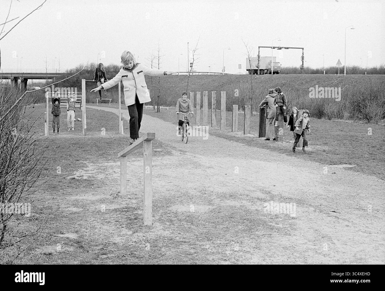 Bambini che giocano intorno all'area di jogging, 01-01-1973 Whizgle News, Dutch Desk, Paesi Bassi, 1950-2000 Un bambino si bilancia con cura su una trave di legno, mentre altri nelle vicinanze si divertono a varie attività, tra cui ciclismo e passeggiate, in una spaziosa area all'aperto con sentieri erbosi e basse strutture in legno. Foto Stock