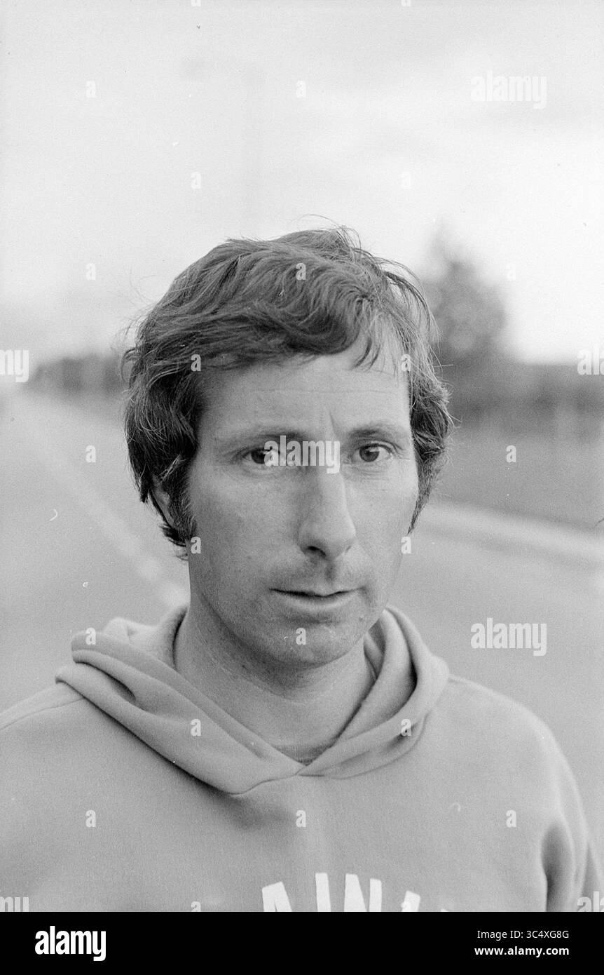 Un allenatore di calcio Whizgle News, Dutch Desk, Paesi Bassi, 1950-2000 Un uomo si trova in primo piano con un'espressione seria, indossando una felpa con cappuccio. Lo sfondo presenta una vista sfocata di una strada e degli alberi, suggerendo un ambiente esterno. Foto Stock