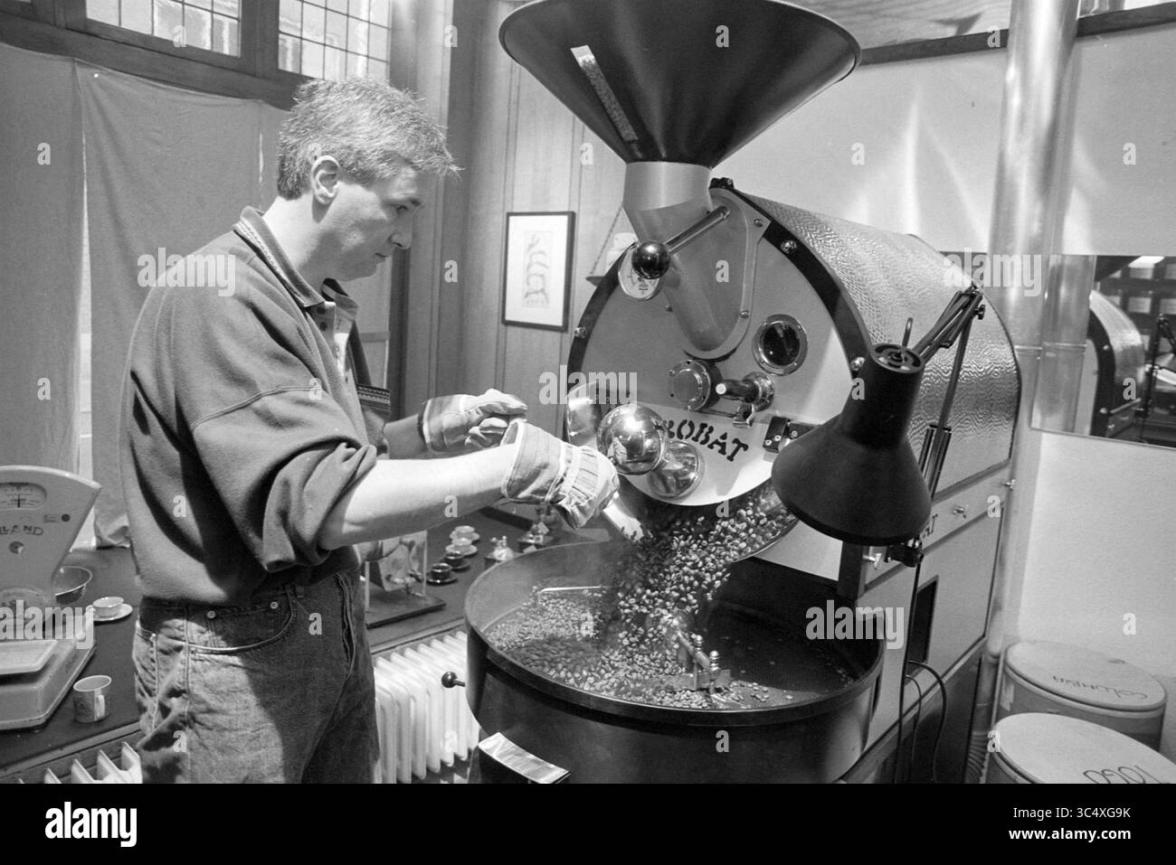 Coffee Roaster de Vries, Haarlem, Paesi Bassi, 07-02-1993 Whizgle News, Dutch Desk, paesi Bassi, 1950-2000 Un barista esperto versa abilmente chicchi di caffè verdi in una grande macchina da tostatura, preparandoli per il processo di tostatura. L'atmosfera calda e invitante del negozio suggerisce la passione per la preparazione della birra perfetta. Foto Stock