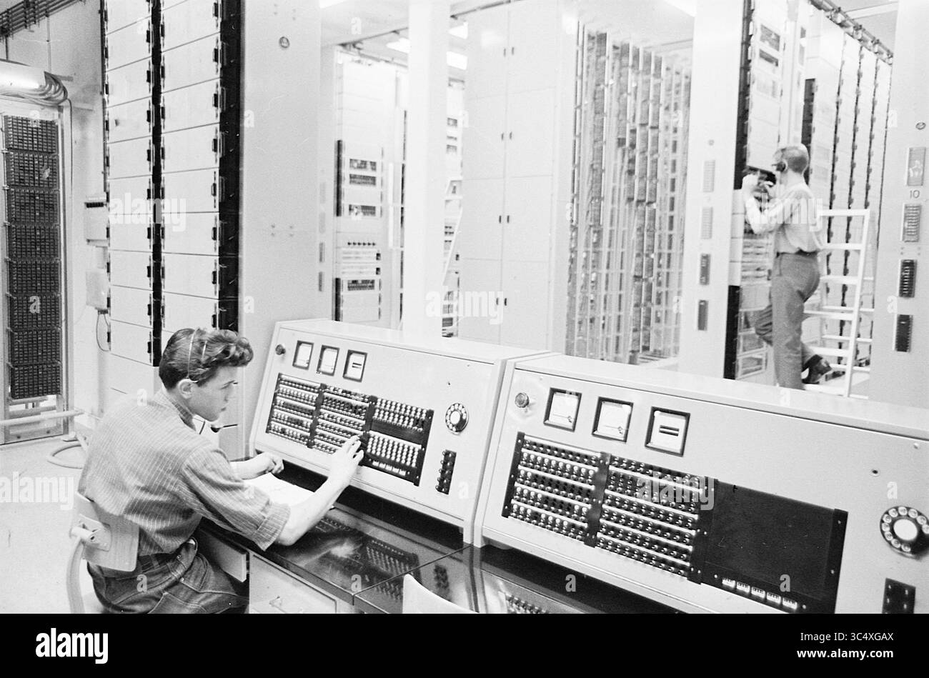 Ufficio centrale di Heemstede, telefono, telefonia, 26-05-1964 Whizgle News, sportello olandese, paesi Bassi, 1950-2000 Un tecnico gestisce una console di computer d'epoca, regolando meticolosamente i quadranti e gli interruttori, mentre un altro individuo cattura la scena sullo sfondo circondata da torreggianti scaffali di cablaggi e apparecchiature, evidenziando un momento di progresso tecnologico in un'epoca passata. Foto Stock