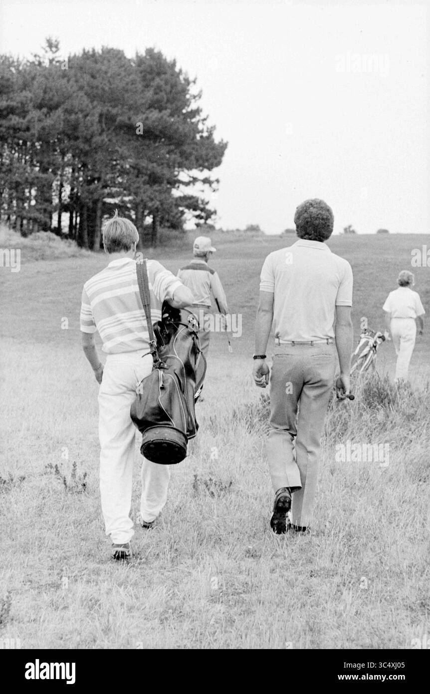 Caddy at Zandvoort golf club, golf, golf, Zandvoort, 20-07-1983 Whizgle News, Dutch Desk, Paesi Bassi, 1950-2000 due persone attraversano un paesaggio erboso, trasportando mazze da golf e chiacchierando piacevolmente. Sullo sfondo, un gruppo di golfisti si fa strada su un dolce pendio, incorniciato da alberi e dalla vasta campagna. L'atmosfera suggerisce una giornata rilassata sul campo, piena di cameratismo e divertimento all'aperto. Foto Stock