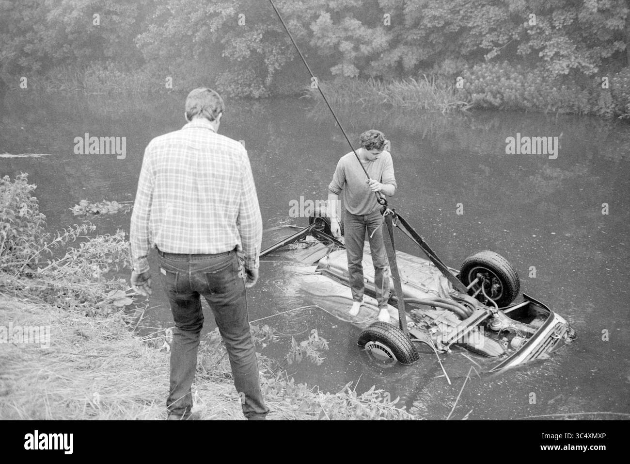 Car in water, 27-07-1985 Whizgle News, Dutch Desk, Paesi Bassi, 1950-2000 due persone stanno accanto a un corpo d'acqua, uno che osserva mentre l'altro lavora per tirare un'auto parzialmente sommersa dalle profondità. La scena trasmette un mix di intrigo e preoccupazione, incorniciata da una lussureggiante vegetazione sullo sfondo. Foto Stock